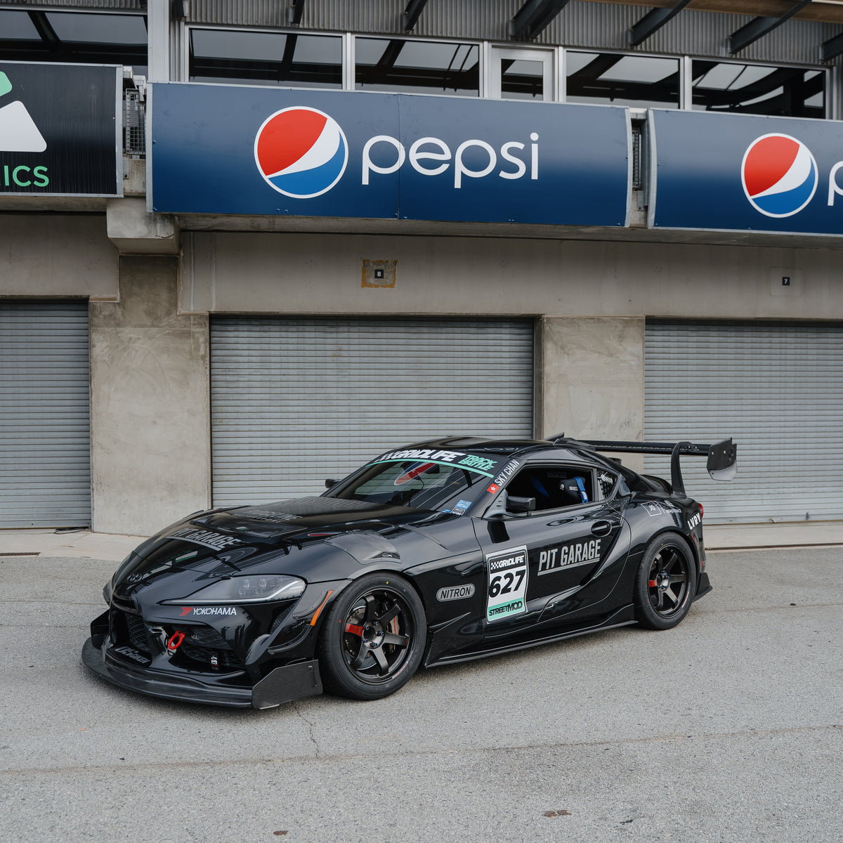 Front three-quarter view of a black Toyota GR Supra race car with the RM Engineering GT4+ front splitter and rear wing outside the pit garages