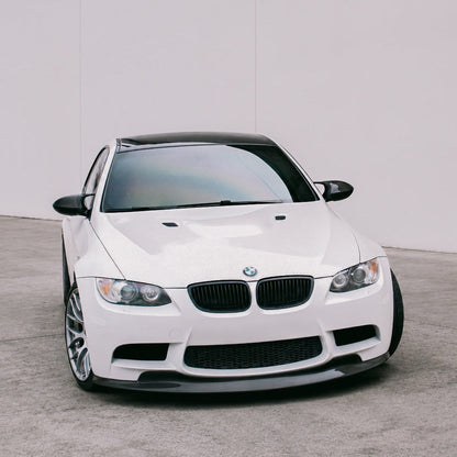 AlpineX Dynamics GTS Style Carbon Fiber Front Lip installed on a BMW E9X M3, shown from a slight side angle highlighting its glossy carbon fiber finish and aggressive aerodynamic profile.