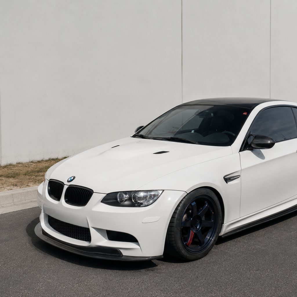 White BMW E9X M3 equipped with the AlpineX Dynamics GTS Style Carbon Fiber Front Lip, shown from the side on a driveway beside a warehouse.