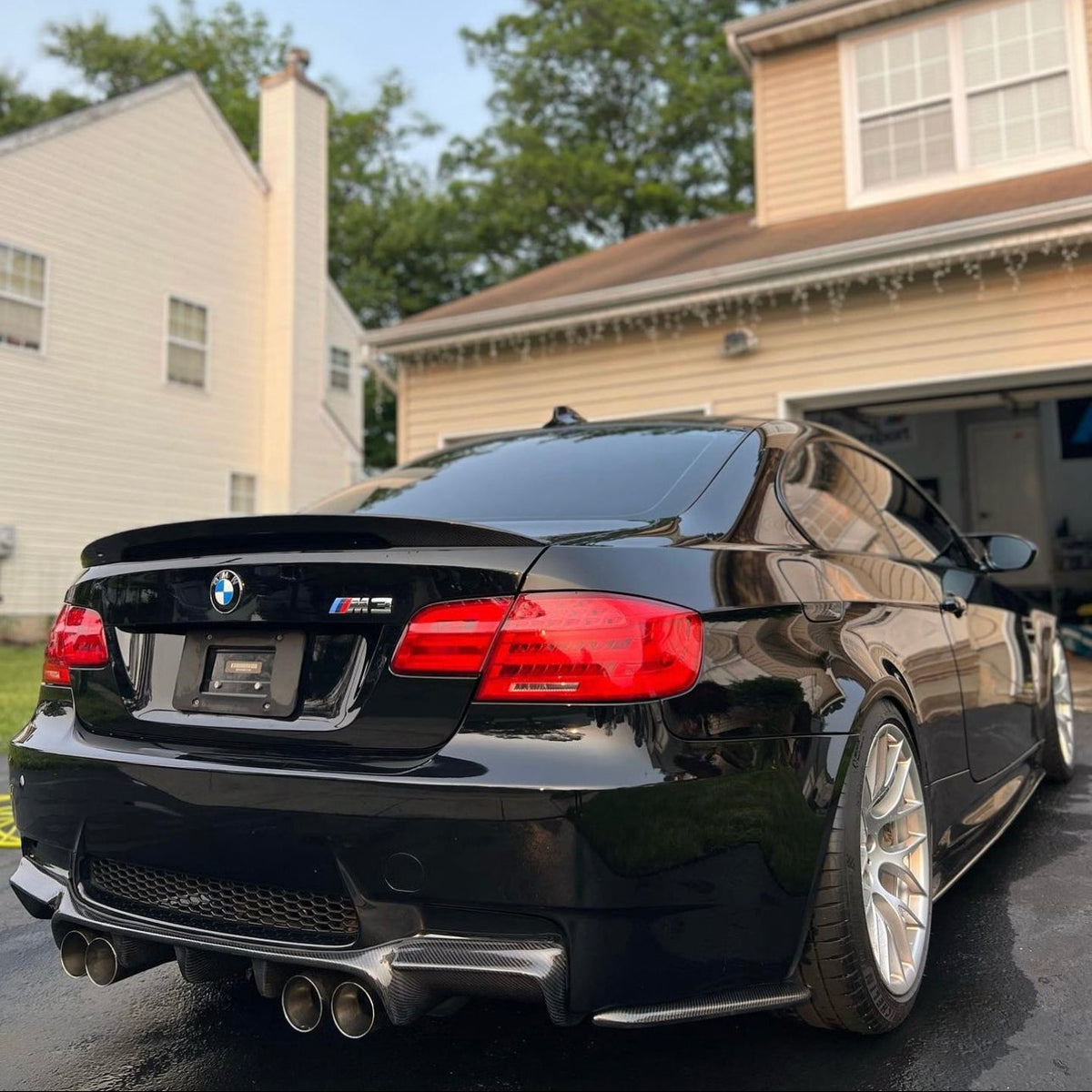 AlpineX Dynamics GTS style carbon fiber rear diffuser for BMW E92/E93 M3 installed on a black coupe, shown from the rear three-quarter angle with quad exhaust tips