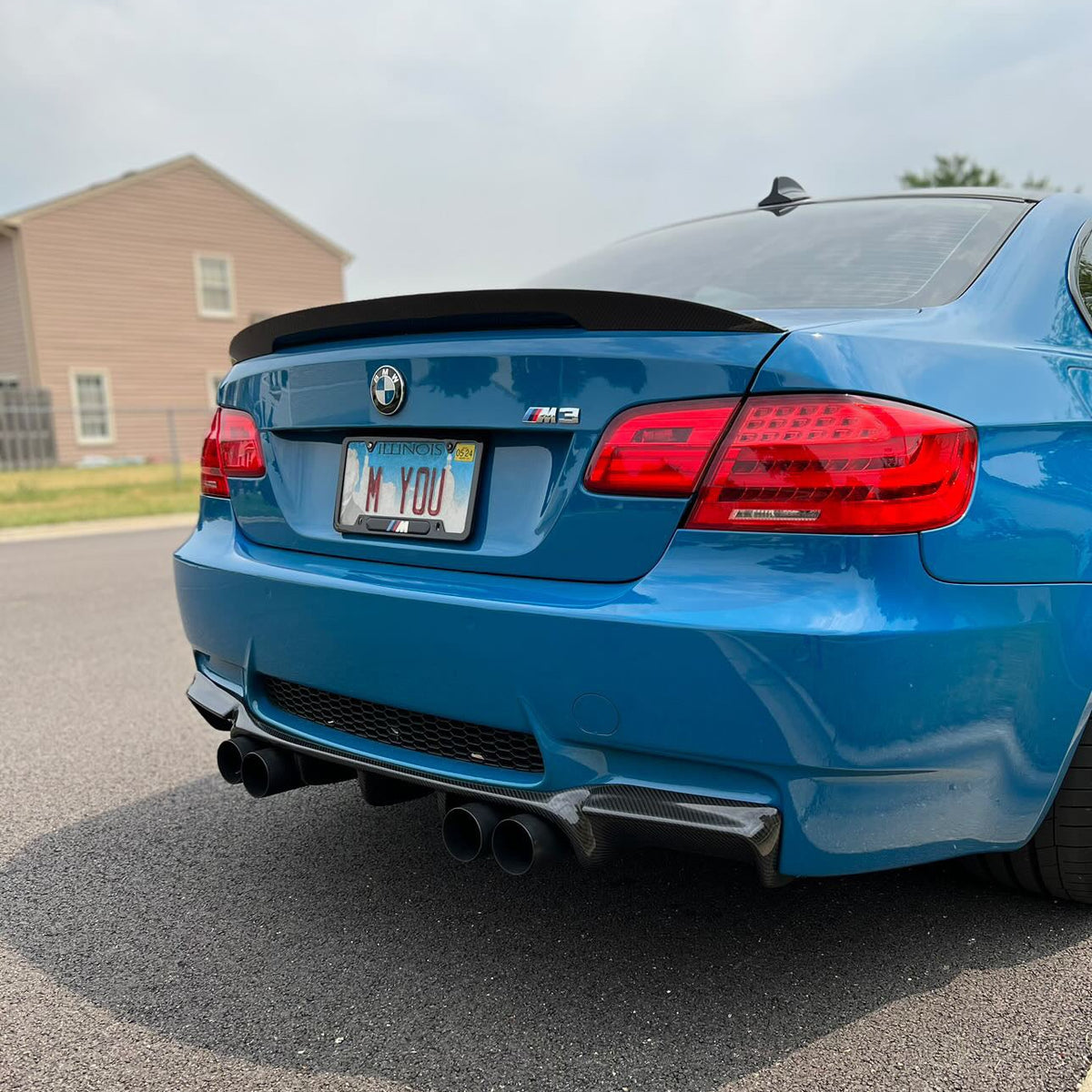 AlpineX Dynamics GTS style carbon fiber rear diffuser for BMW E92/E93 M3 installed on a blue coupe, showing its sculpted fins beneath the rear bumper and quad exhaust tips