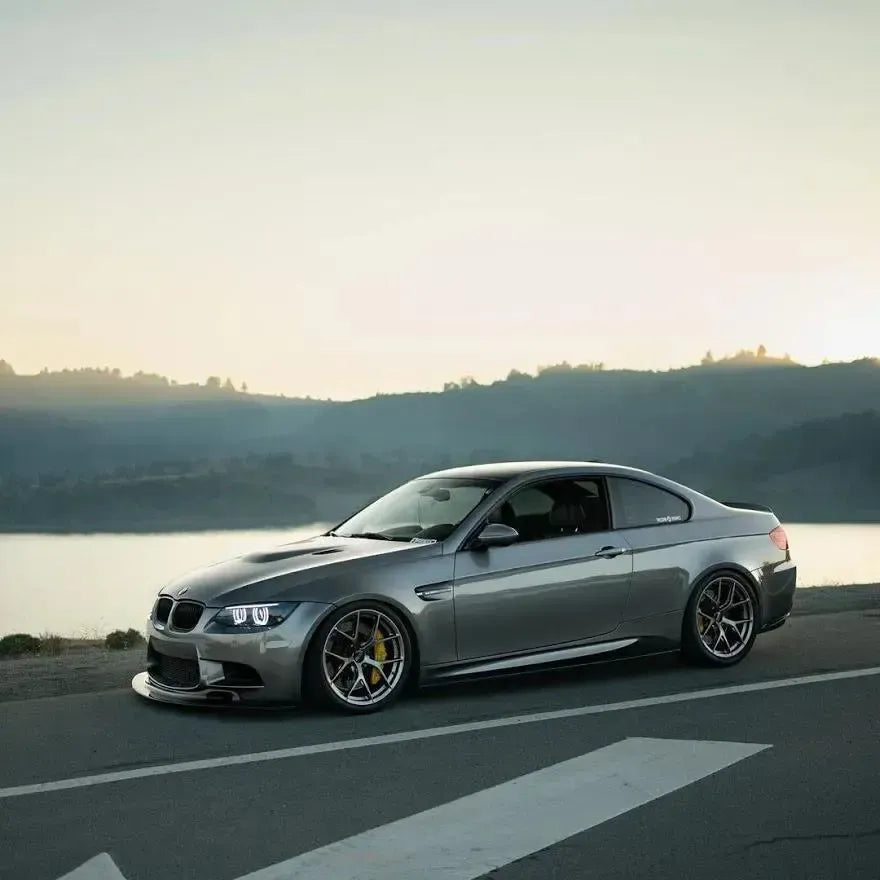 Side three-quarter view of a gray BMW E92 M3 with AlpineX Dynamics MP style carbon fiber side skirts parked beside a lake