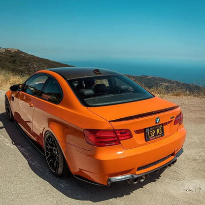 Rear three-quarter view of an orange BMW E92 M3 with AlpineX Dynamics MP style carbon fiber side skirts parked at a coastal overlook