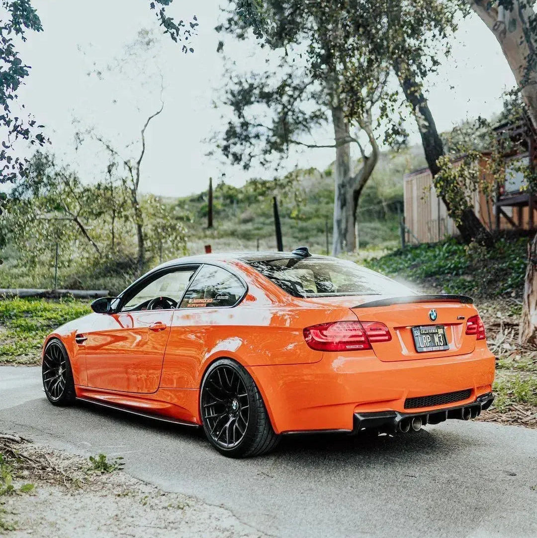 Rear side view of an orange BMW E92 M3 showing AlpineX Dynamics MP style carbon fiber side skirts along the lower body on a tree-lined road