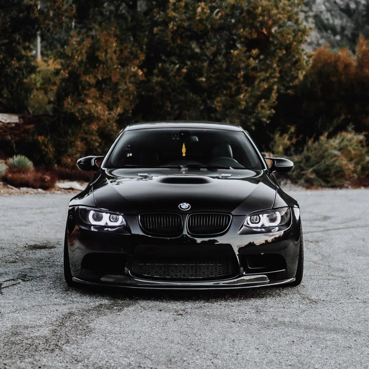 Front view of a black BMW E9X M3 with an AlpineX Dynamics GTS style one-piece carbon fiber front lip, parked on gravel with trees in the background