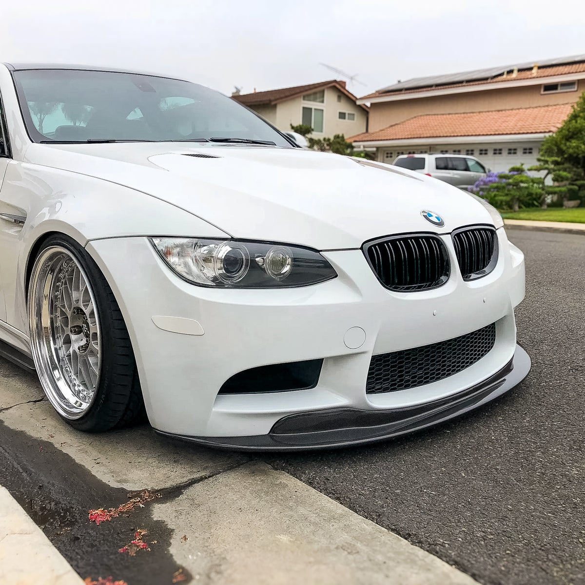 AlpineX Dynamics GTS style one-piece carbon fiber front lip installed on a white BMW E9X M3, shown from a front three-quarter angle beside a residential curb