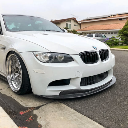 AlpineX Dynamics GTS style one-piece carbon fiber front lip installed on a white BMW E9X M3, shown from a front three-quarter angle beside a residential curb