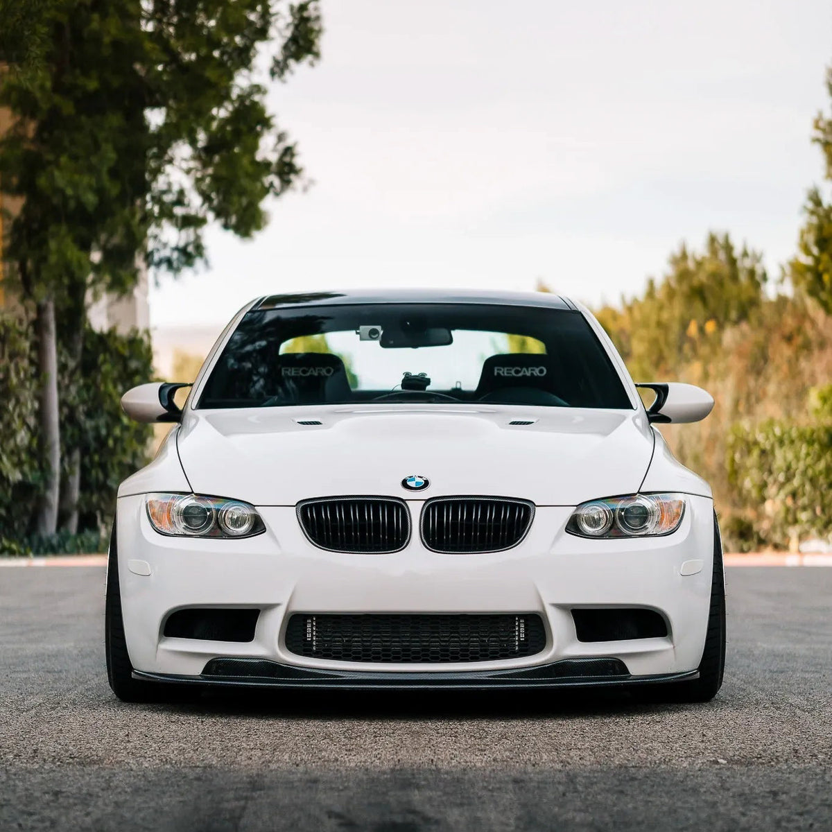 Head-on view of a white BMW E9X M3 with an AlpineX Dynamics GTS style one-piece carbon fiber front lip and black kidney grilles, parked on a tree-lined road