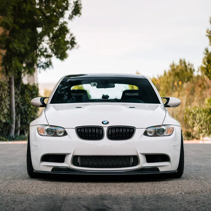 Head-on view of a white BMW E9X M3 with an AlpineX Dynamics GTS style one-piece carbon fiber front lip and black kidney grilles, parked on a tree-lined road