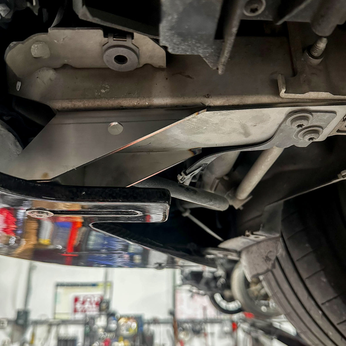 Underbody mounting detail for the Ridgeline Motorsport adjustable carbon fiber front splitter on a BMW F87 M2 OG N55, showing the metal support bracket and mounting bolts
