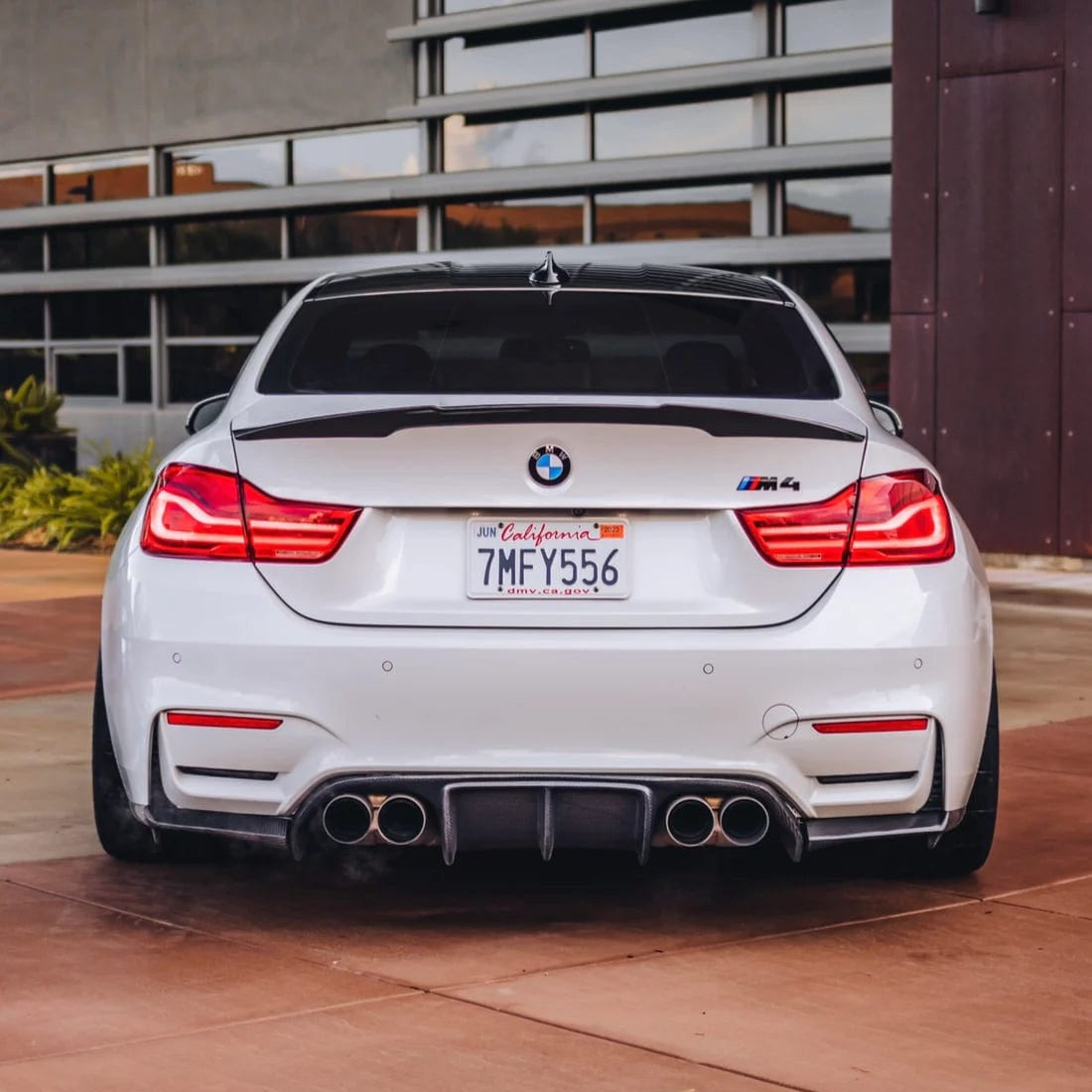 Rear view of a white BMW M4 with the AlpineX Dynamics V style three-piece carbon fiber rear diffuser for F8X M3/M4, showing the central fins, side extensions and quad exhaust tips
