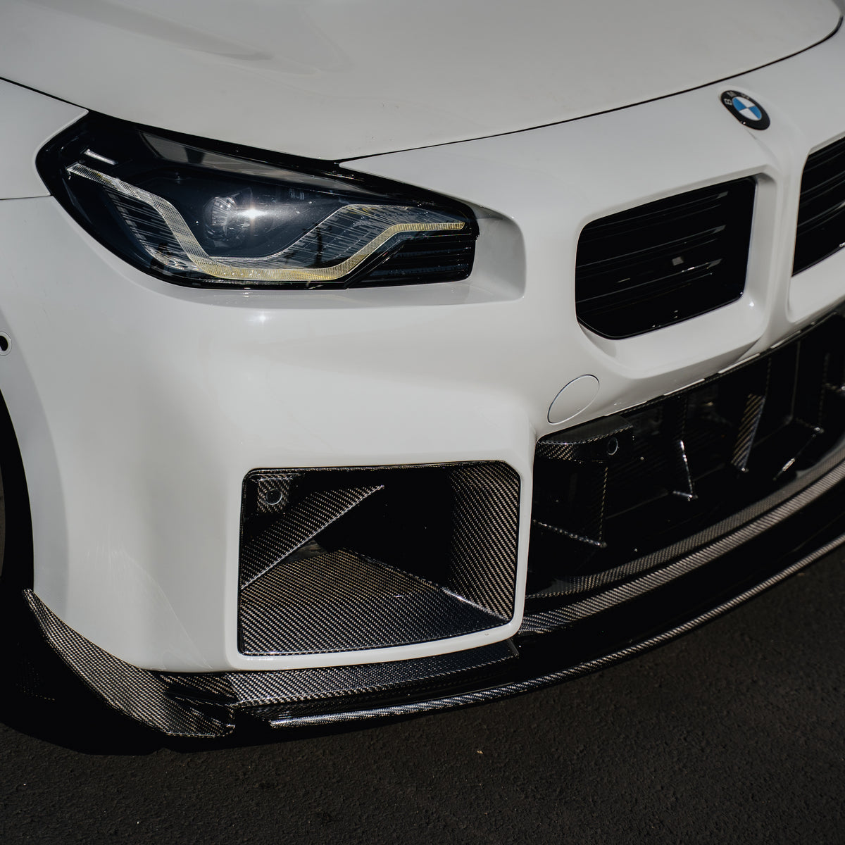 Close-up of the Ridgeline Motorsport adjustable carbon fiber front splitter beneath the front bumper and lower grille of a white BMW G87 M2