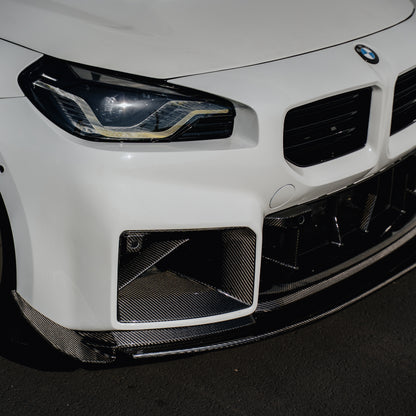 Close-up of the Ridgeline Motorsport adjustable carbon fiber front splitter beneath the front bumper and lower grille of a white BMW G87 M2