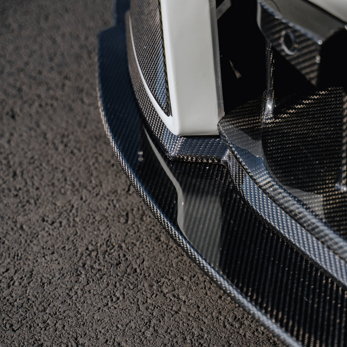 Close-up of the woven carbon fiber surface and leading edge of the Ridgeline Motorsport adjustable front splitter installed beneath a white BMW G87 M2 bumper