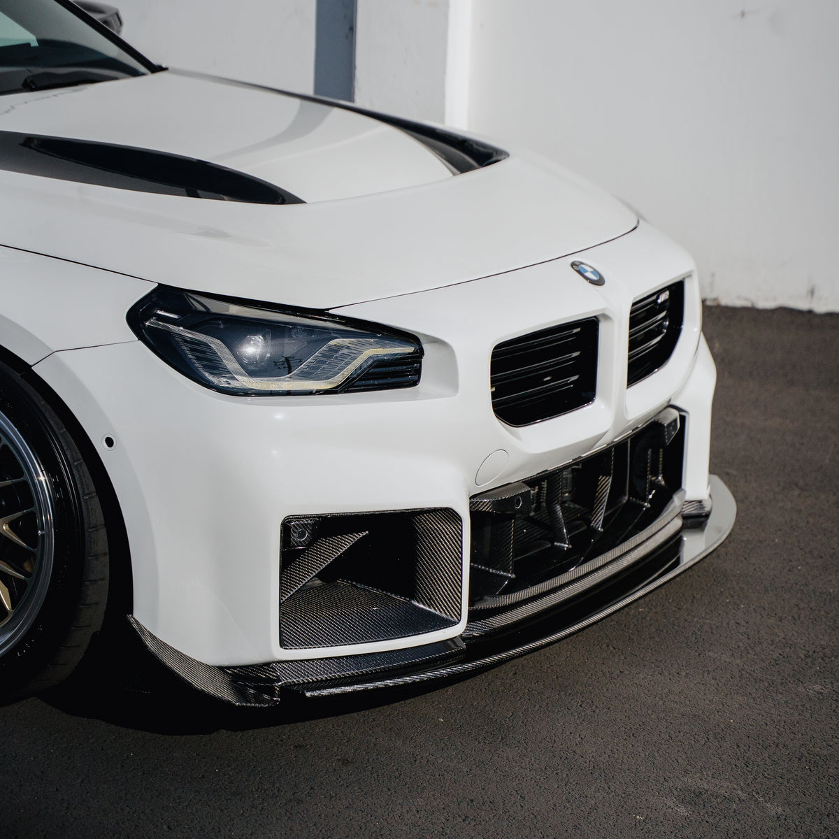 Close-up of the Ridgeline Motorsport adjustable carbon fiber front splitter on a white BMW G87 M2, showing the splitter edge beneath the front bumper and carbon air intakes