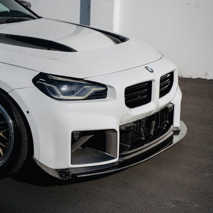 Close-up of the Ridgeline Motorsport adjustable carbon fiber front splitter on a white BMW G87 M2, showing the splitter edge beneath the front bumper and carbon air intakes