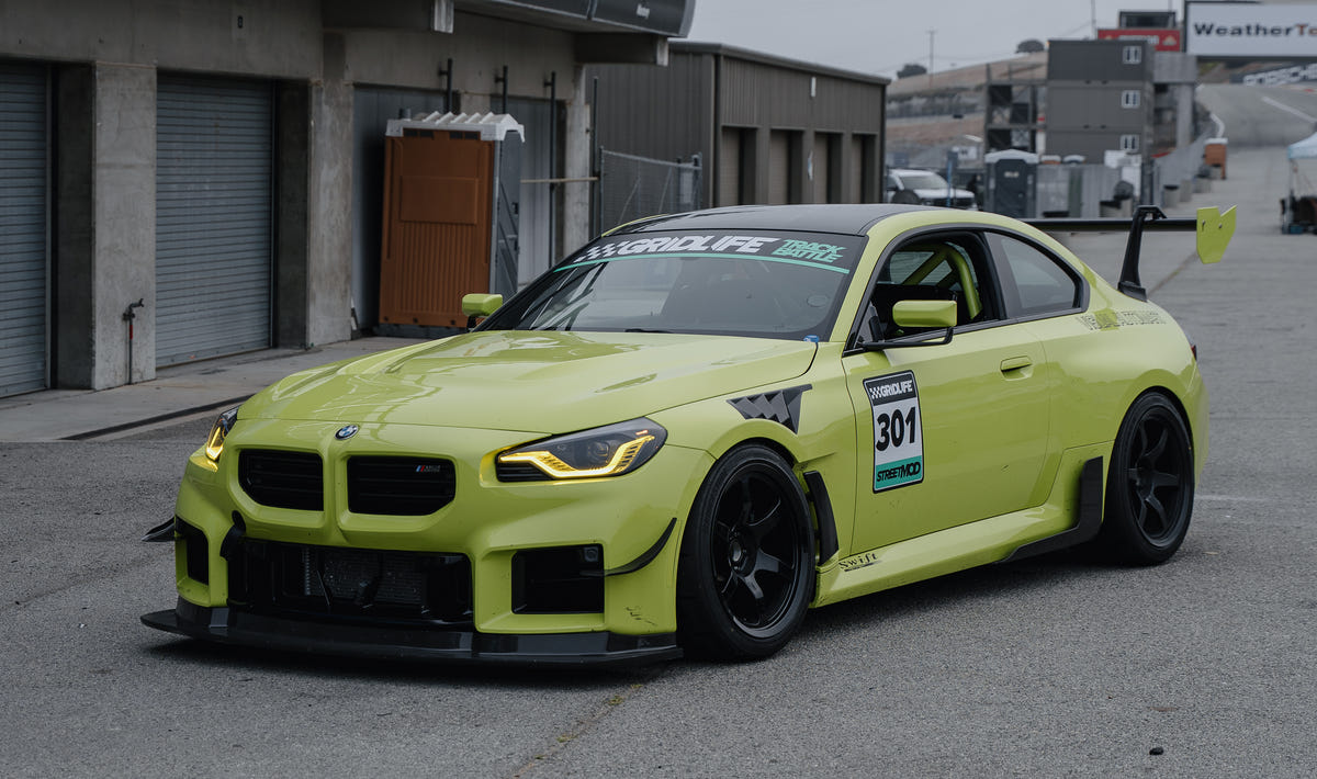Yellow BMW G87 M2 race car number 301 parked in the pit lane, showing its front splitter, bumper canards and large rear wing from a front three-quarter view