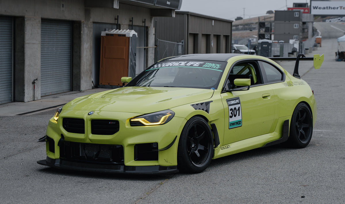 Yellow BMW G87 M2 race car number 301 parked in the pit lane, showing its front splitter, bumper canards and large rear wing from a front three-quarter view