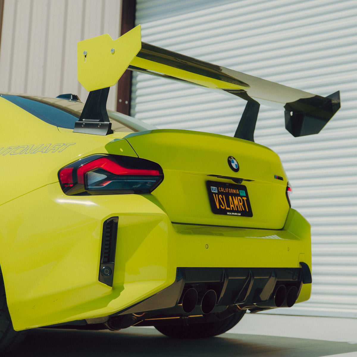 Low rear view of a Sao Paulo Yellow BMW G87 M2 showing the GT4+ carbon fiber wing, swan-neck uprights and trunk mounts