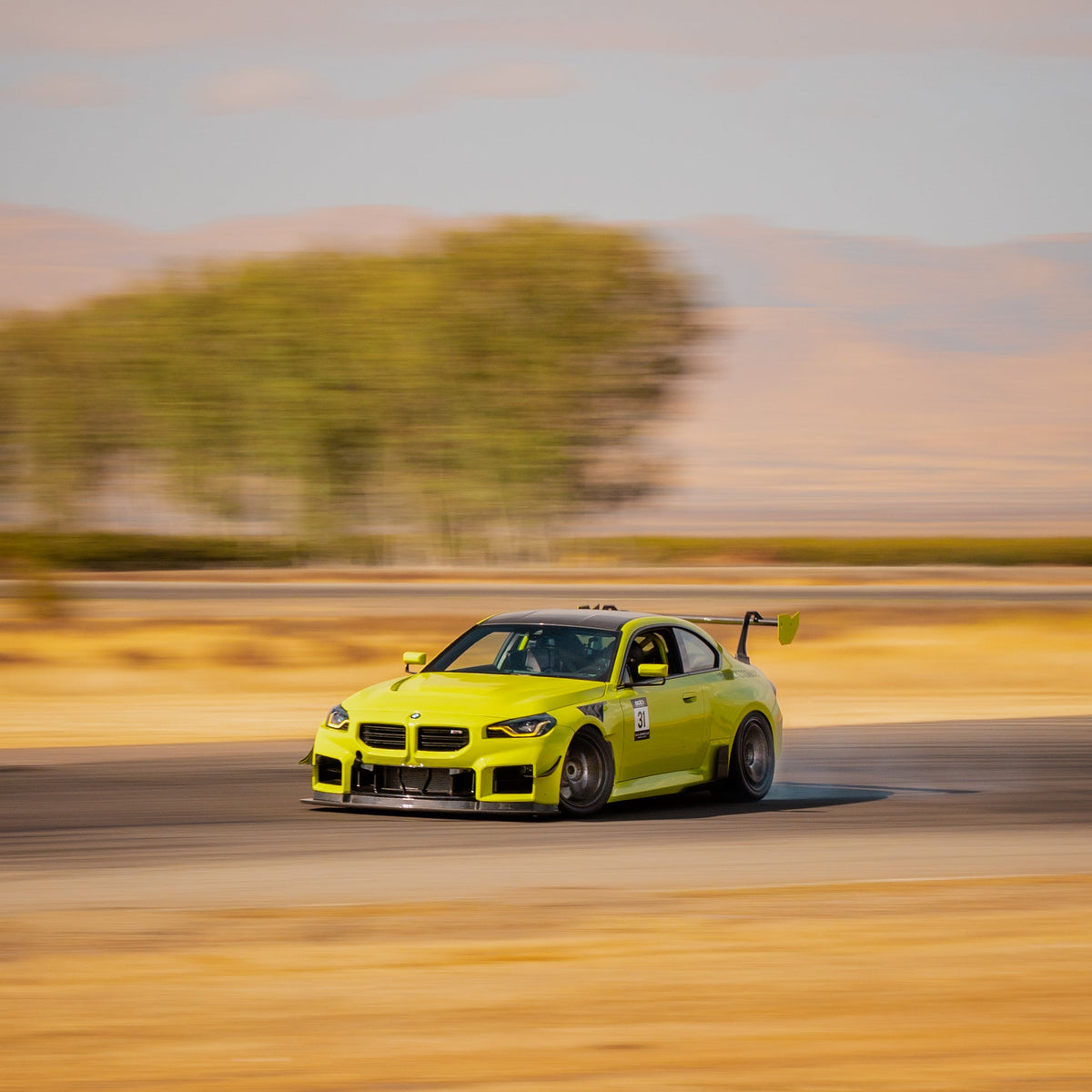 Front three-quarter track view of a Sao Paulo Yellow BMW G87 M2 equipped with the GT4+ rear wing