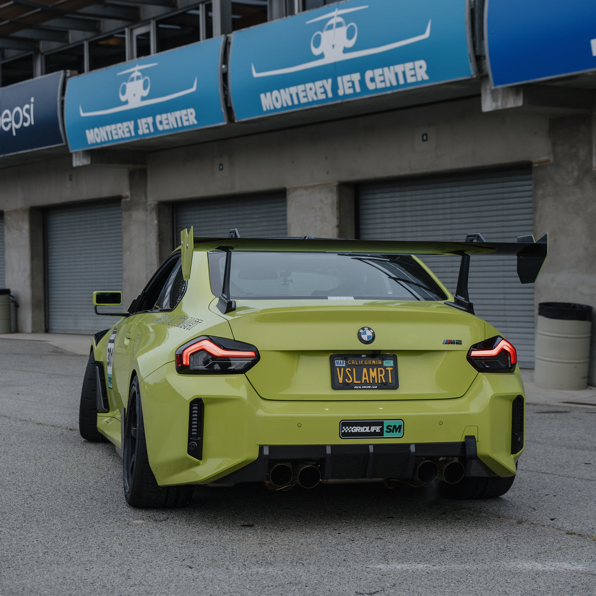 Rear view of a Sao Paulo Yellow BMW G87 M2 showing the full span and swan-neck uprights of the RM Engineering GT4+ rear wing outside the pit garages