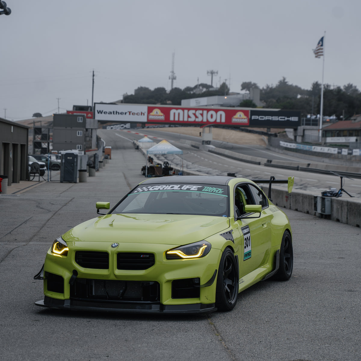 Front three-quarter view of a Sao Paulo Yellow BMW G87 M2 with the RM Engineering GT4+ swan-neck rear wing parked in the pit lane