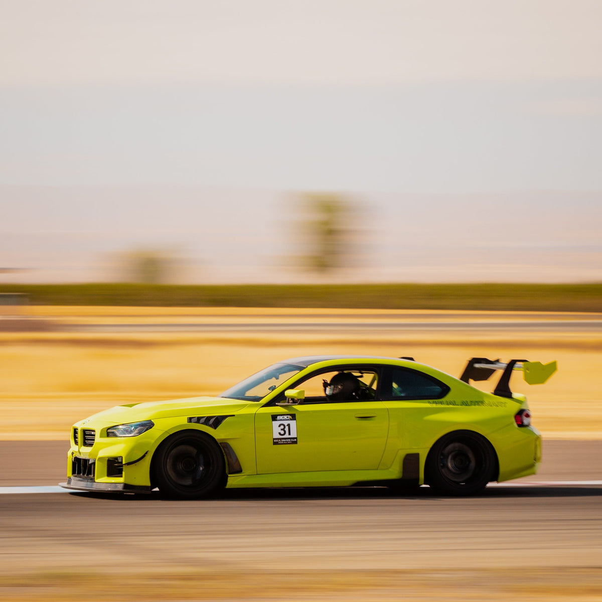 Close track-side view of a Sao Paulo Yellow BMW G87 M2 with the GT4+ swan-neck rear wing