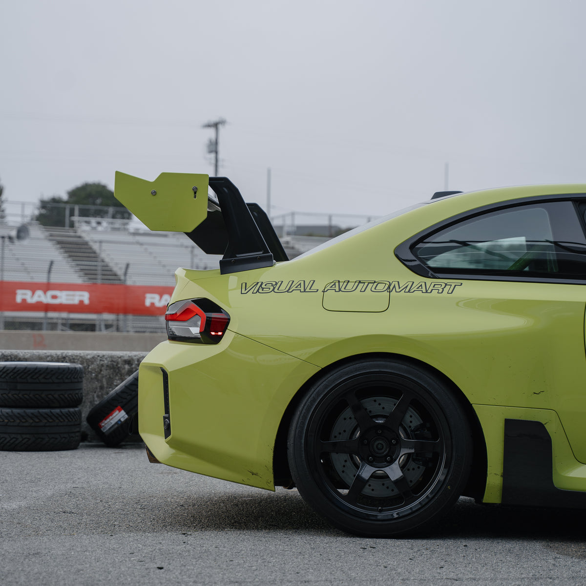 Trackside rear side detail of a Sao Paulo Yellow BMW G87 M2 showing the RM Engineering GT4+ swan-neck wing, yellow endplate and trunk mount