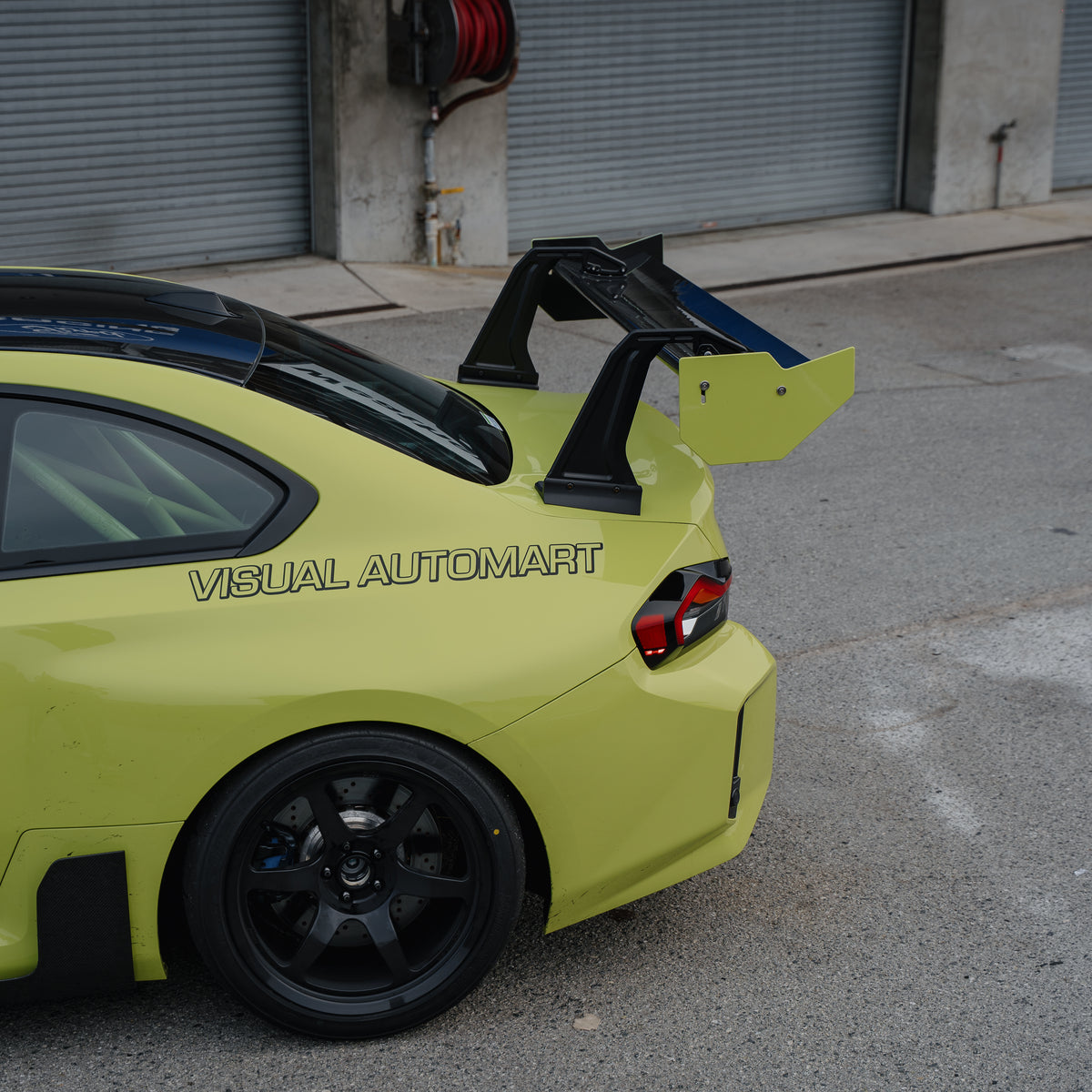 Upper rear-quarter detail of the RM Engineering GT4+ swan-neck wing with yellow endplates on a Sao Paulo Yellow BMW G87 M2 outside the pit garages