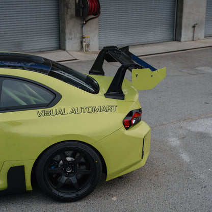 Upper rear-quarter detail of the RM Engineering GT4+ swan-neck wing with yellow endplates on a Sao Paulo Yellow BMW G87 M2 outside the pit garages