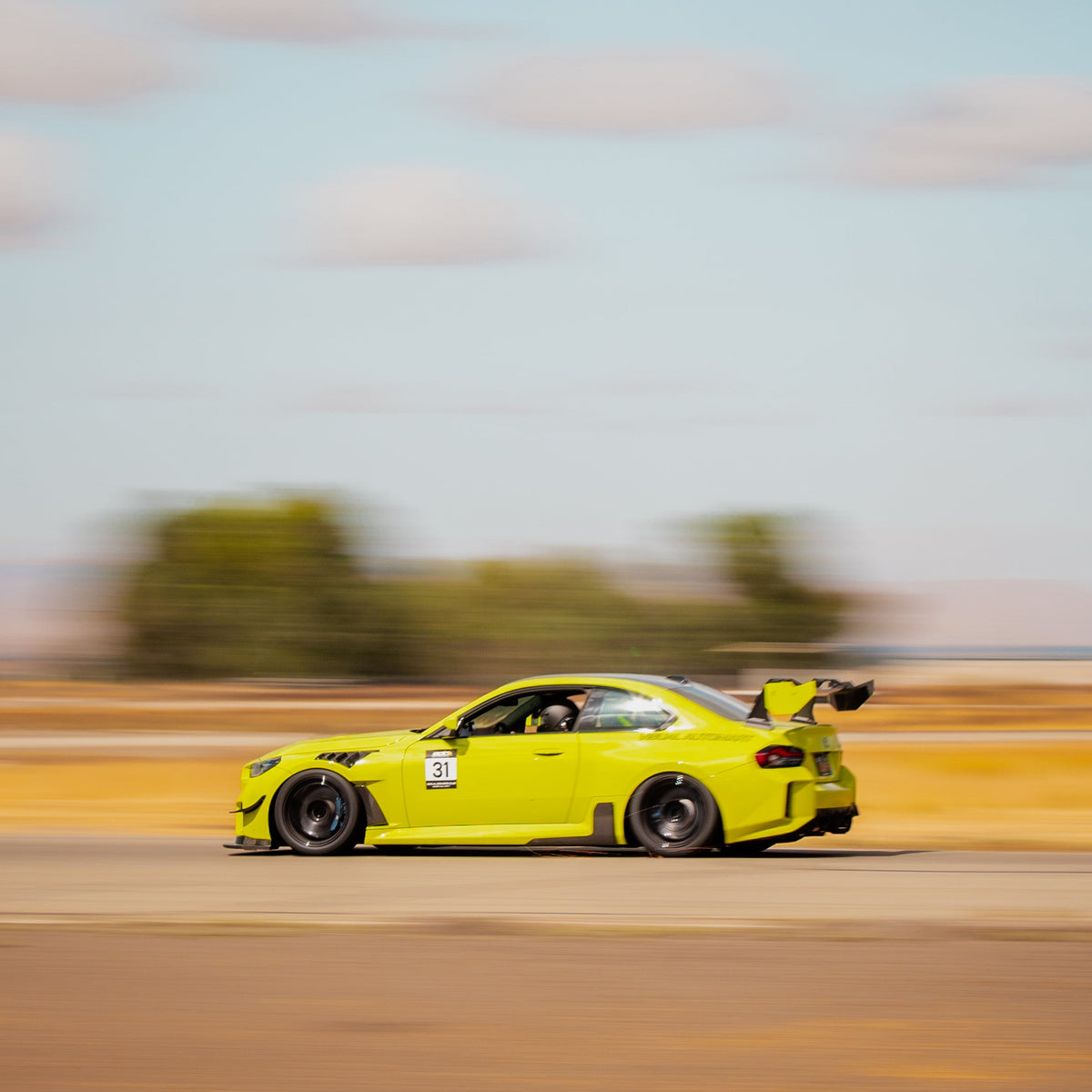 Rear three-quarter motion view of a BMW G87 M2 showing the GT4+ rear wing during track driving