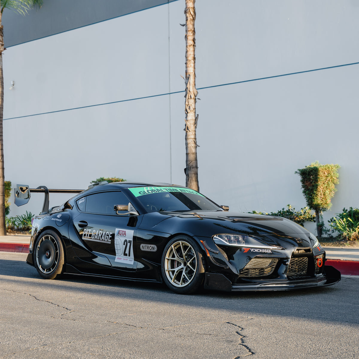 Black Toyota GR Supra A90/A91 race car with the RM Engineering GT4+ rear wing, photographed from a front three-quarter angle outside a gray building