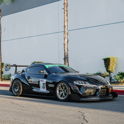Black Toyota GR Supra A90/A91 race car with the RM Engineering GT4+ rear wing, photographed from a front three-quarter angle outside a gray building