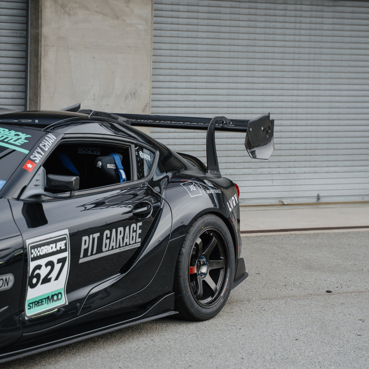 Side detail of the RM Engineering GT4+ rear wing, swan-neck upright and endplate on a black Toyota GR Supra race car outside a pit garage