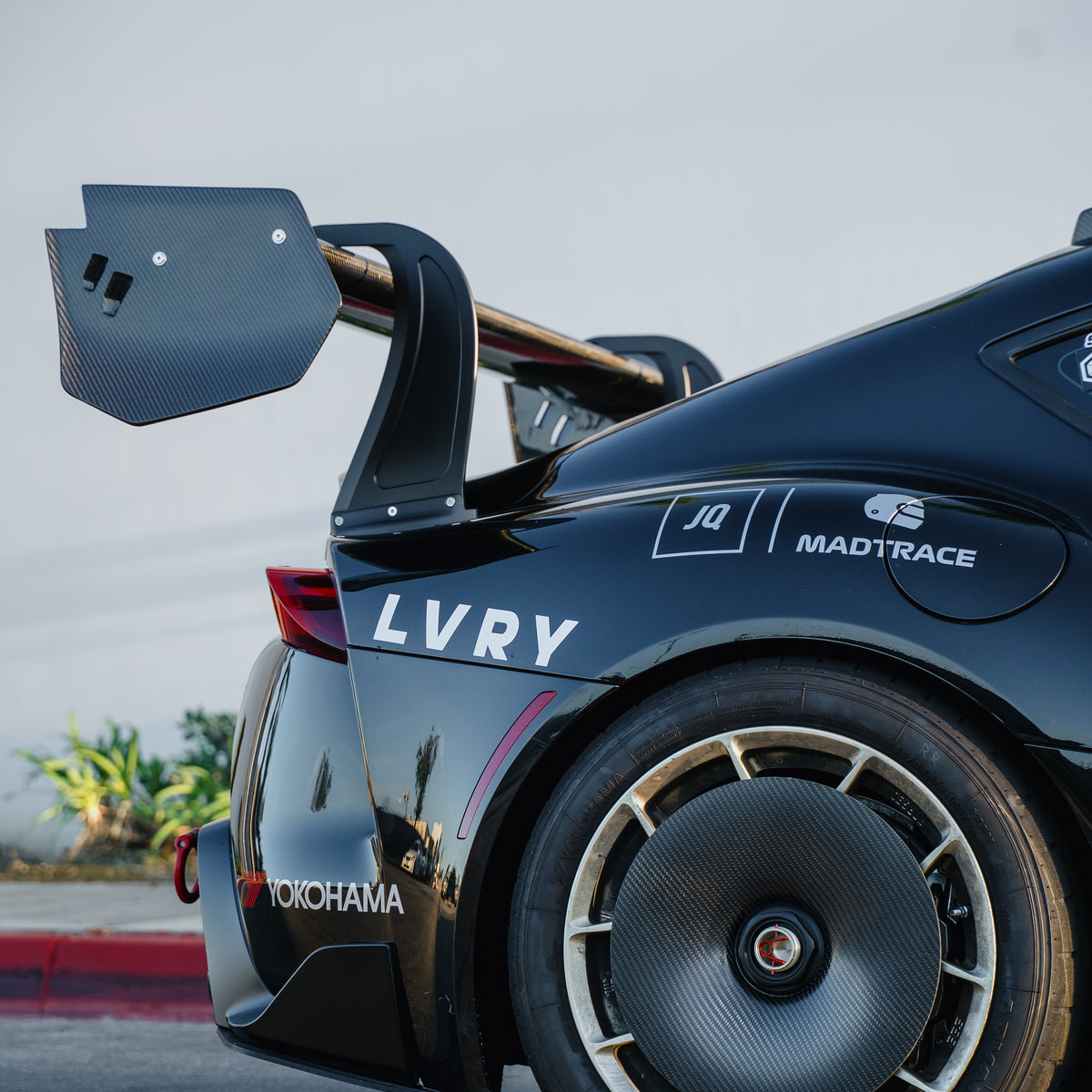 Close-up of the RM Engineering GT4+ rear wing on a black Toyota GR Supra A90/A91, showing the carbon fiber endplate and upright above the rear wheel and fender