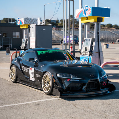 Black Toyota GR Supra with the RM Engineering GT4+ rear wing for A90 and A91 models, shown from a front three-quarter angle beside race fuel pumps