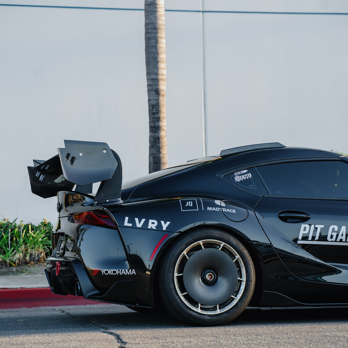 Rear three-quarter detail of a black Toyota GR Supra A90/A91 with the RM Engineering GT4+ rear wing, showing its carbon fiber endplates above the rear fender and taillight