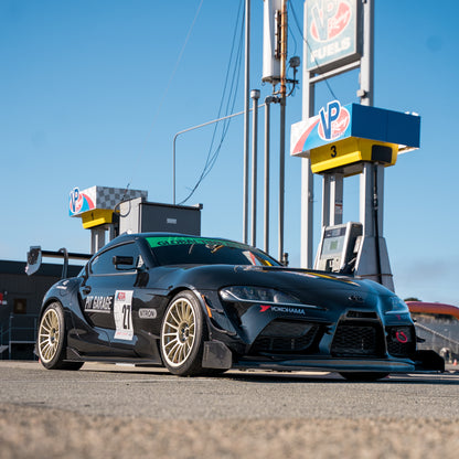 Low-angle front three-quarter view of a black Toyota GR Supra with RM Engineering GT4+ front splitter and rear wing, parked beside race fuel pumps under a blue sky