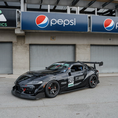Front three-quarter view of a black Toyota GR Supra race car with the RM Engineering GT4+ front splitter and rear wing outside the pit garages