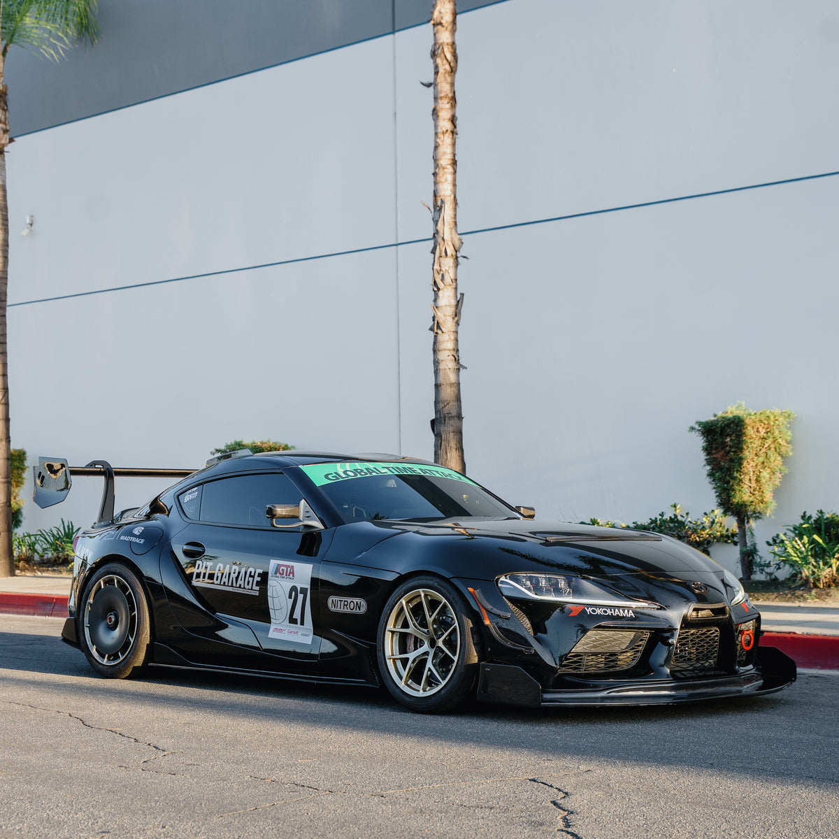Black Toyota GR Supra A90 race car parked outside a building with the RM Engineering GT4+ front splitter kit, shown in a full-car front three-quarter view