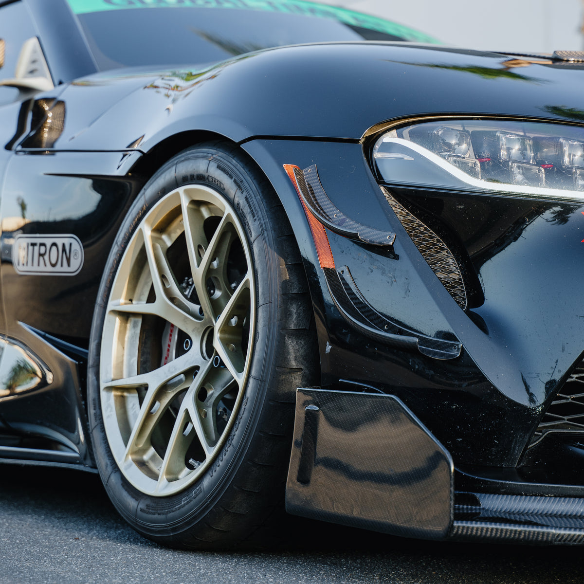 Close-up of the RM Engineering GT4+ front splitter endplate on a black Toyota GR Supra A90, with carbon fiber bumper canards and a gold front wheel
