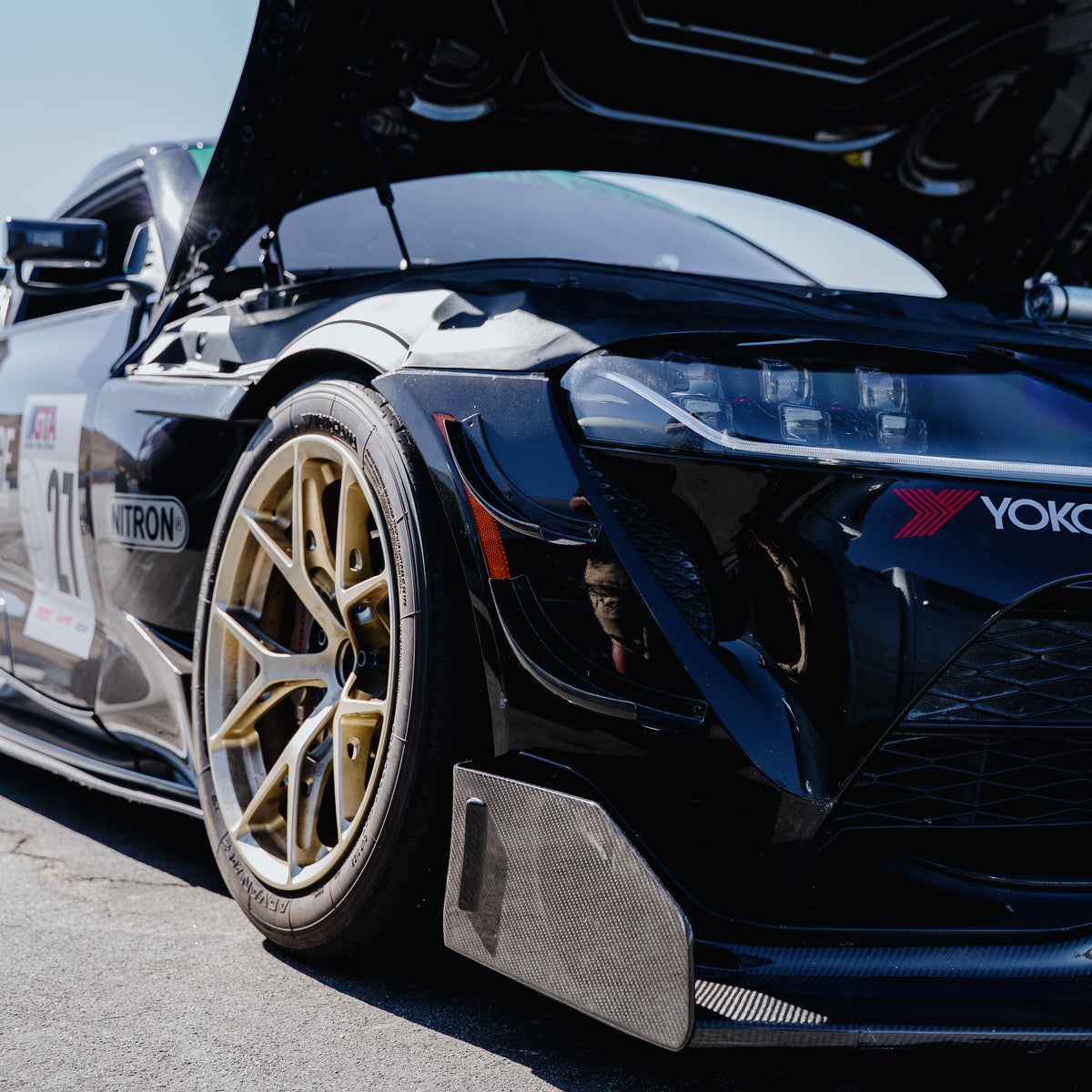 Close-up of the RM Engineering GT4+ carbon fiber front splitter and endplate on a black Toyota GR Supra A90 with the hood open, showing the bumper canards and gold wheel