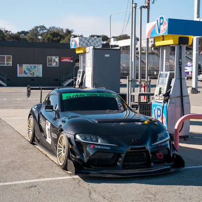 Black Toyota GR Supra A90 fitted with the RM Engineering GT4+ front splitter kit, parked beside race fuel pumps with its carbon splitter and endplates visible